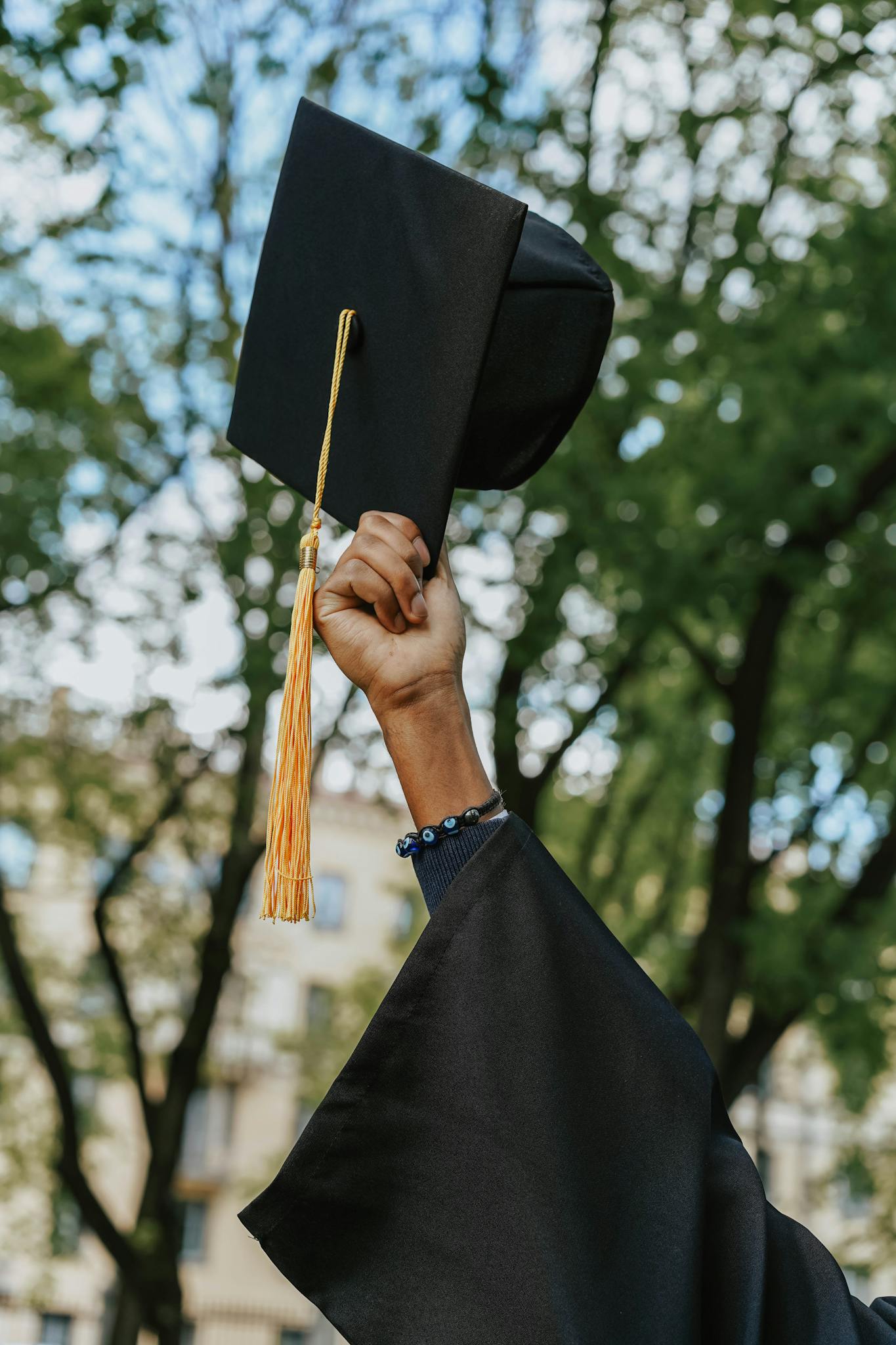 A close-up of a hand raising a graduation cap in celebration among trees.